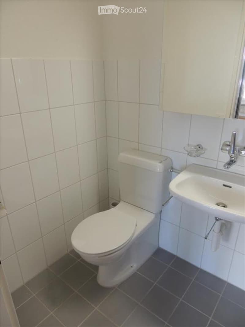 A small, clean bathroom with white tiled walls, a toilet, a sink with a mirror, and a white cabinet above the sink.
