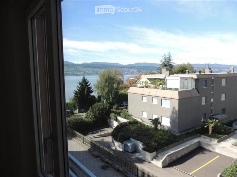 View from a window showing a modern multi-story building with a balcony, surrounded by greenery and overlooking a lake and distant mountains.