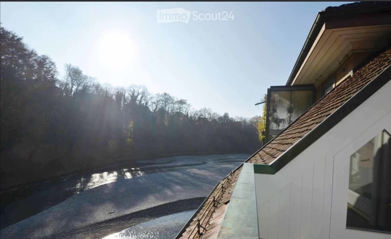 A modern house with a tiled roof and white exterior, situated near a river with trees in the background.