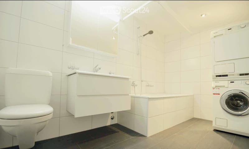 Modern bathroom with white tiles, a toilet, a sink with a mirror, a bathtub with a shower, and a stacked washing machine and dryer.