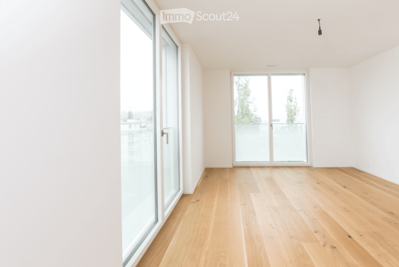 Empty room with light wooden flooring, white walls, and large glass windows and doors leading to a balcony or outdoor area.