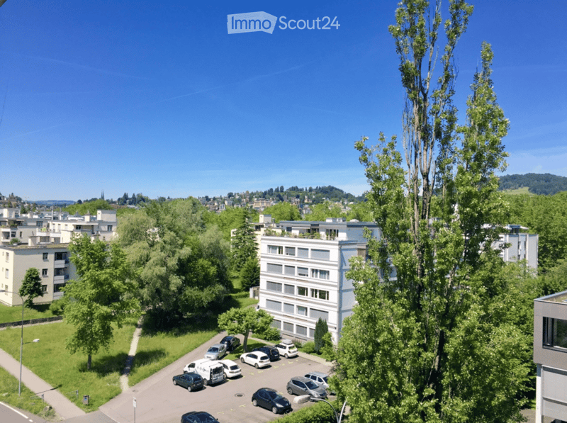 Aerial view of a residential area with multiple apartment buildings, a parking lot with several cars, and surrounding greenery.