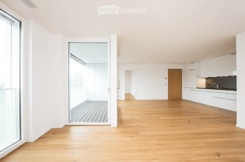 Bright, open-plan space with light wooden flooring, white walls, and large glass windows. The area includes a kitchen with white cabinets and a dark countertop, and a wooden door leading to another room.