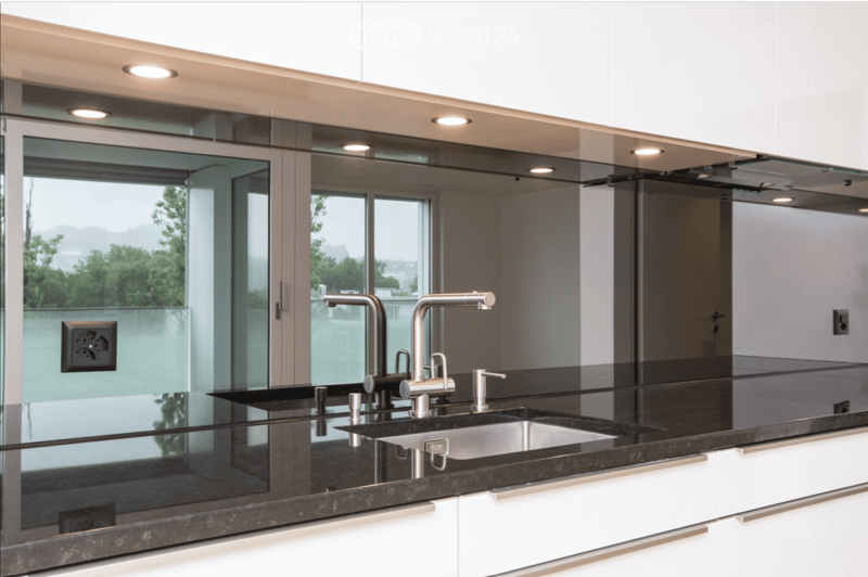 Modern kitchen with white cabinets, black granite countertops, and a double sink with stainless steel faucets. Large windows offer a view of greenery outside.