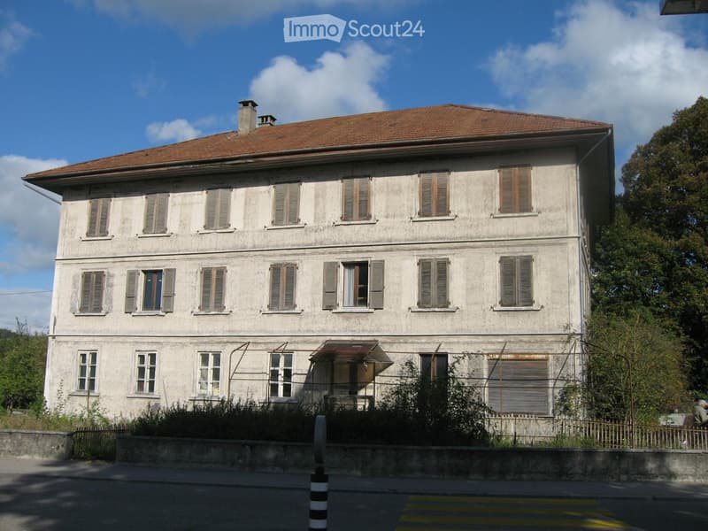 Two-story residential building with beige facade, multiple windows with shutters, and a small awning over the entrance.