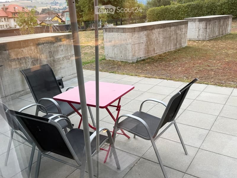 Balcony with tiled floor, pink table, and four black chairs arranged around it. Concrete walls and a grassy area are visible in the background.