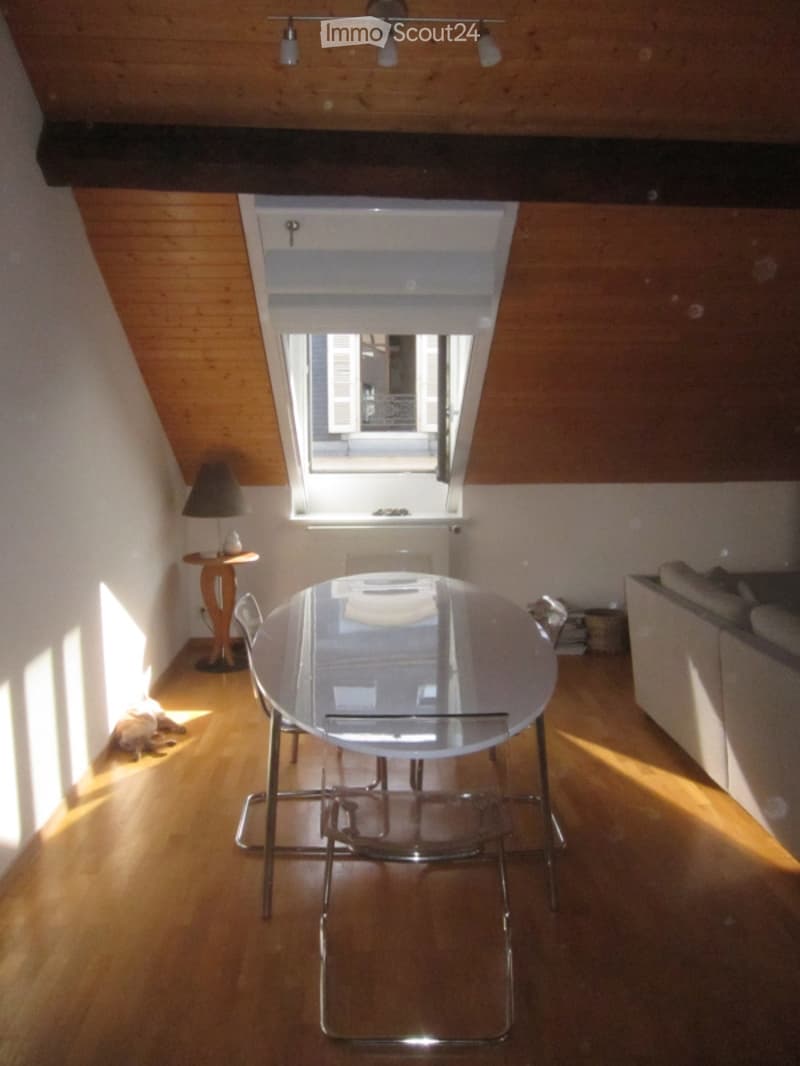 A modern, well-lit room with wooden flooring and exposed wooden beams. Features a sleek white dining table with chairs, a small side table with a lamp, and a comfortable sofa. The room has large windows and a skylight, allowing ample natural light.