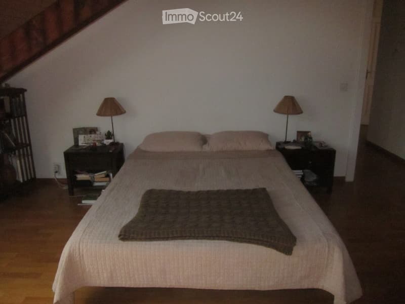 Cozy bedroom with wooden floor, slanted ceiling, bed with pillows and blanket, nightstands with lamps, books, and picture frames.