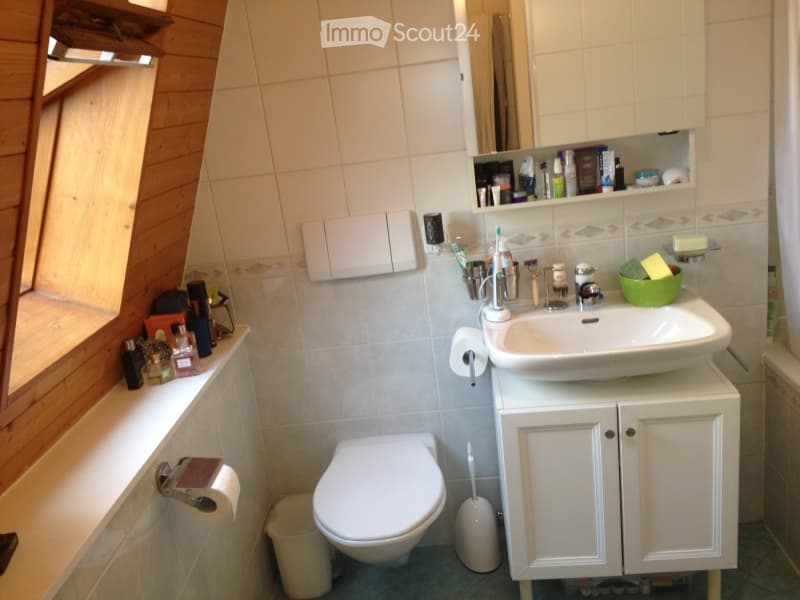 A compact bathroom with a white sink, toilet, and cabinet. The walls are tiled, and there is a window with wooden shutters. Various toiletries and a green bowl are visible on the sink counter.