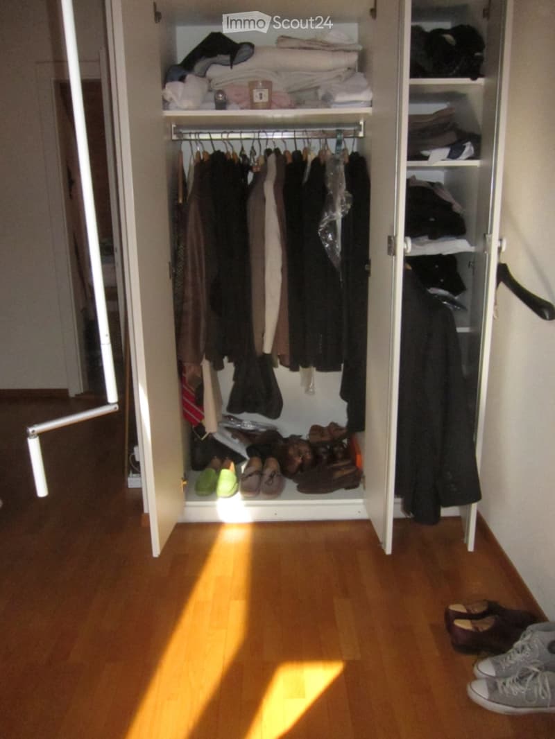 Open wardrobe with clothes, shoes, and folded items, set against a wooden floor and white walls.