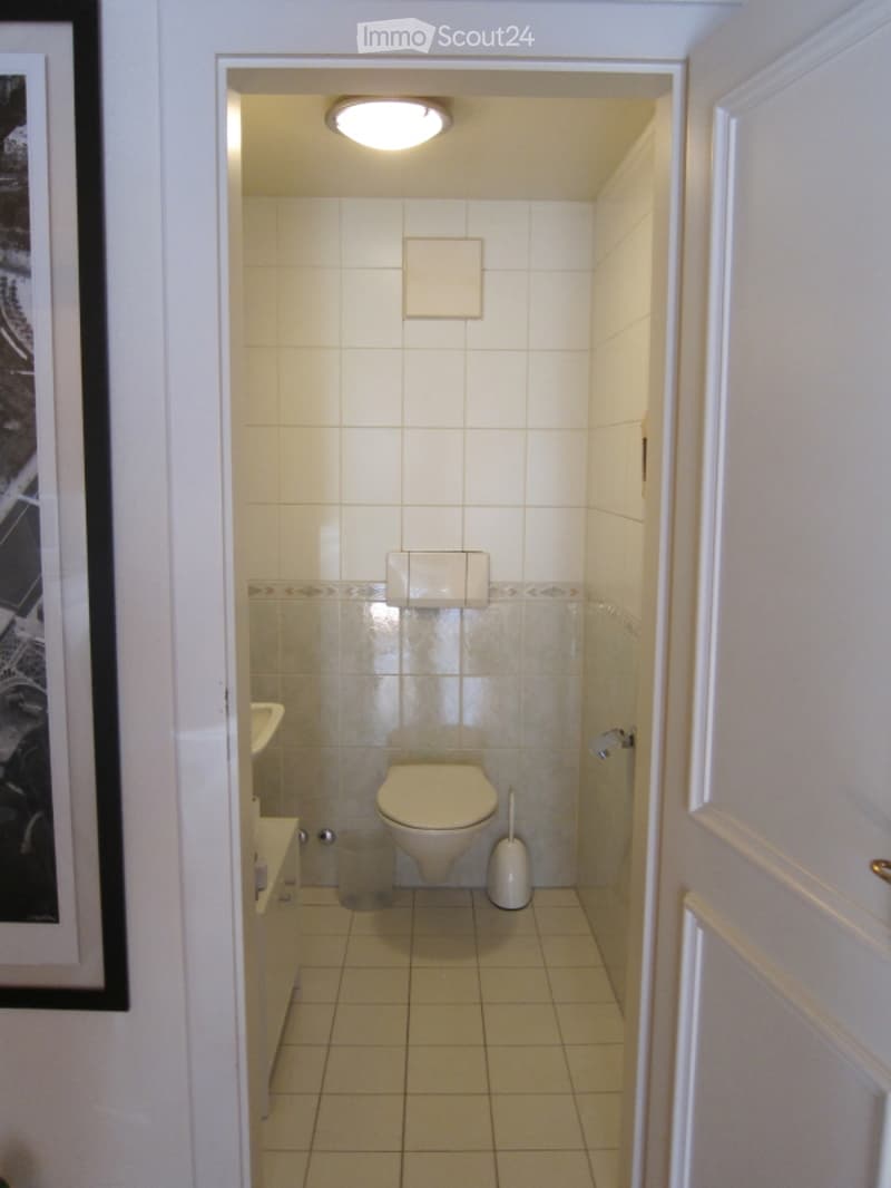 Small, clean bathroom with white tiled walls and floor, a toilet, a sink, and a toilet brush holder.