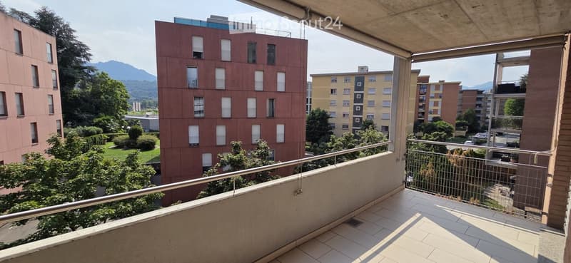Balcony with tiled floor, metal railing, overlooking a residential area with buildings, trees, and distant mountains.