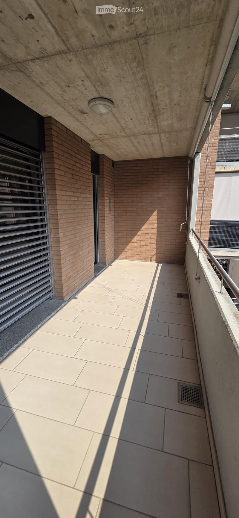 A modern, clean corridor with tiled flooring, brick walls, and a concrete ceiling. Features include a metal railing on one side and a light fixture on the ceiling.