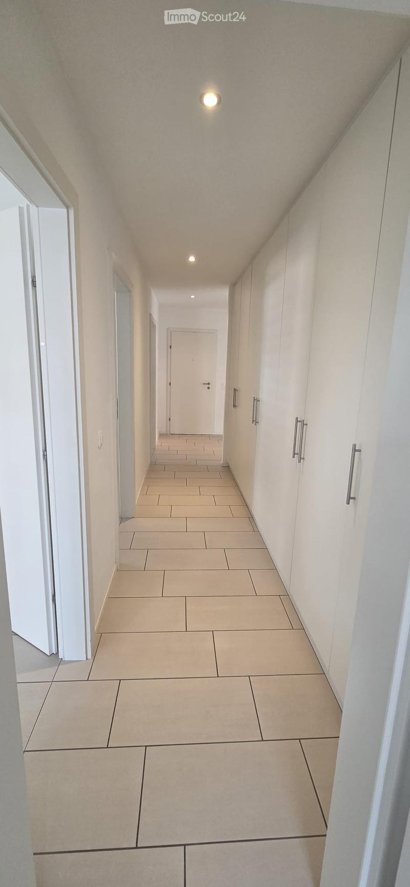 Clean, modern hallway with beige tiled flooring, white walls, and recessed lighting. Features built-in white cabinets with silver handles on one side and several doors leading to other rooms.