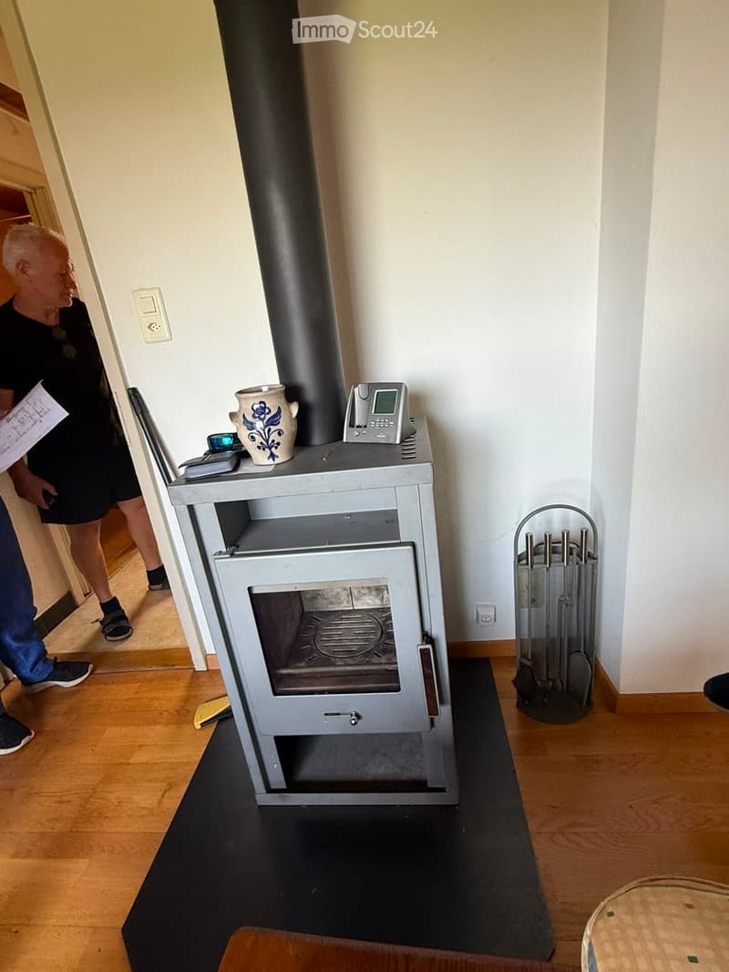 A modern wood-burning stove with a decorative vase and phone on top, surrounded by a wooden floor and white walls.