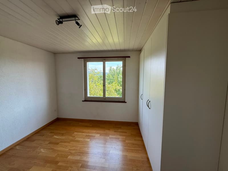 Empty room with wooden flooring, white walls, and a large window offering a view of greenery. The ceiling has built-in lighting fixtures.