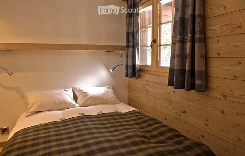 Cozy bedroom with a wooden wall, a bed with white pillows and a plaid blanket, a window with dark curtains, and a wall-mounted lamp providing soft lighting.
