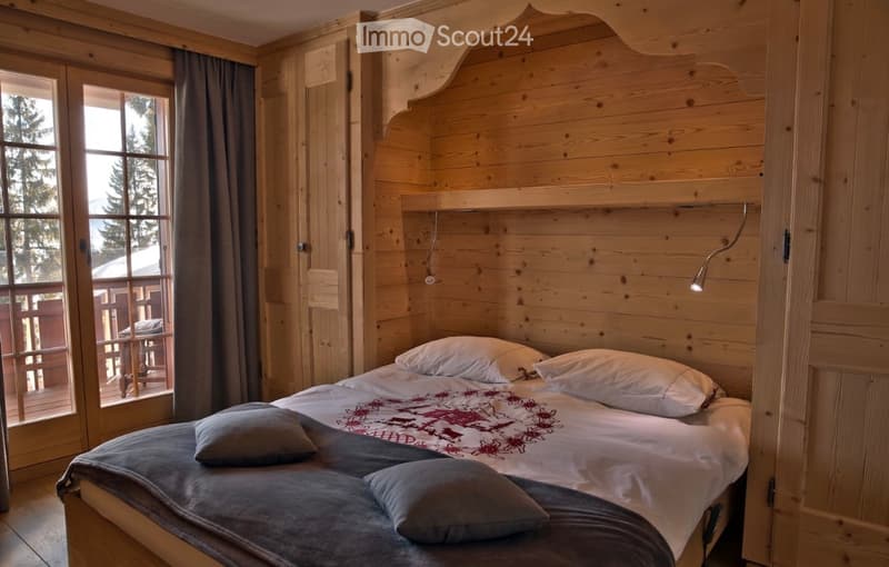Cozy wooden bedroom with a large bed, pillows, and a nightstand lamp. Large windows offer a scenic view of the outdoors.