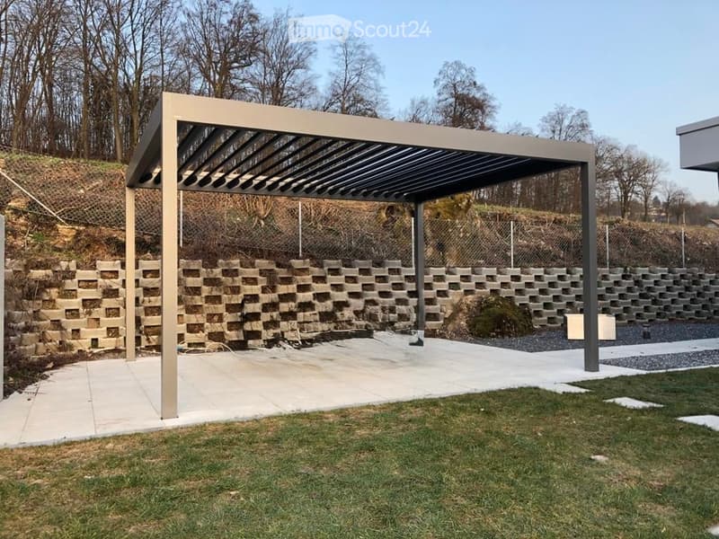 Modern outdoor patio with a pergola, stone retaining wall, and gravel pathway, surrounded by grass and trees.