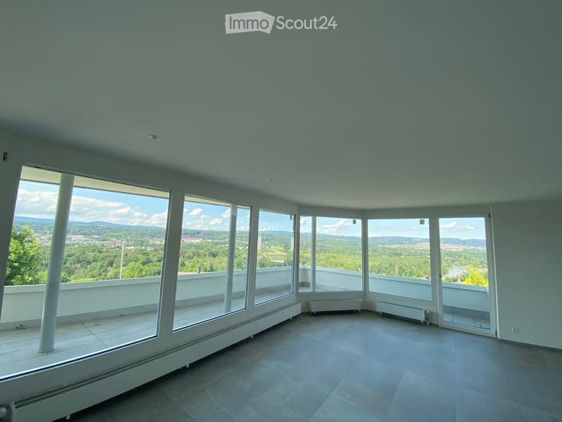Bright, empty room with large windows offering a scenic view of a green landscape and distant cityscape. The room features a tiled floor and white walls.