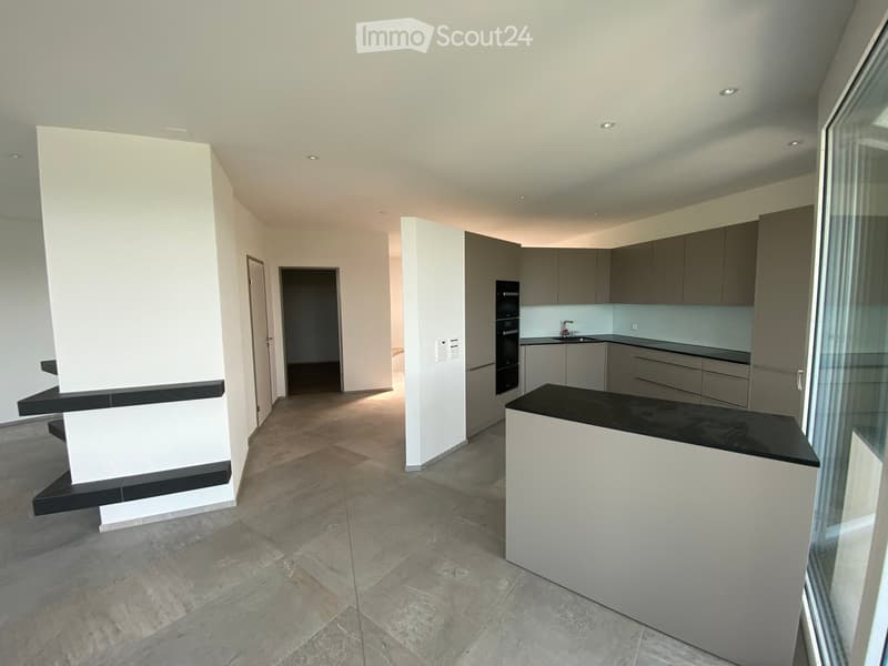 Modern, unfurnished kitchen with sleek grey cabinets, a black countertop, and a built-in oven. The space features large windows and a tiled floor.
