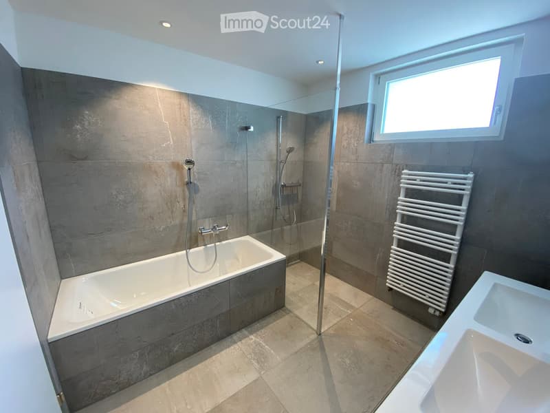 Modern bathroom with a bathtub, sink, radiator, and glass-enclosed shower area. Features grey tiled walls and a large window providing natural light.