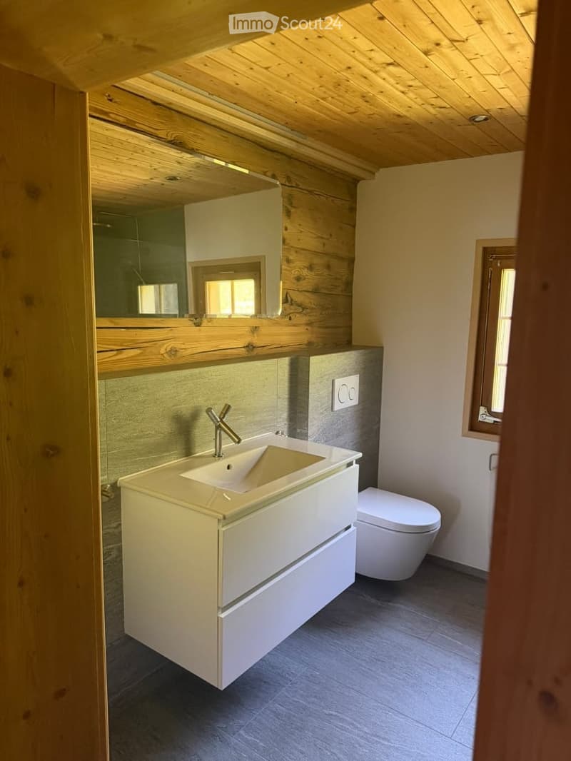 Modern bathroom with a white vanity, sleek sink, chrome faucet, and a large mirror above. The room features wooden accents and a grey tiled floor.