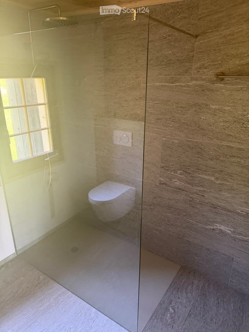 Modern bathroom with a glass shower enclosure, a toilet, and a showerhead. The walls are tiled in a light, textured pattern, and the floor is tiled in a light wood finish. A small window provides natural light.