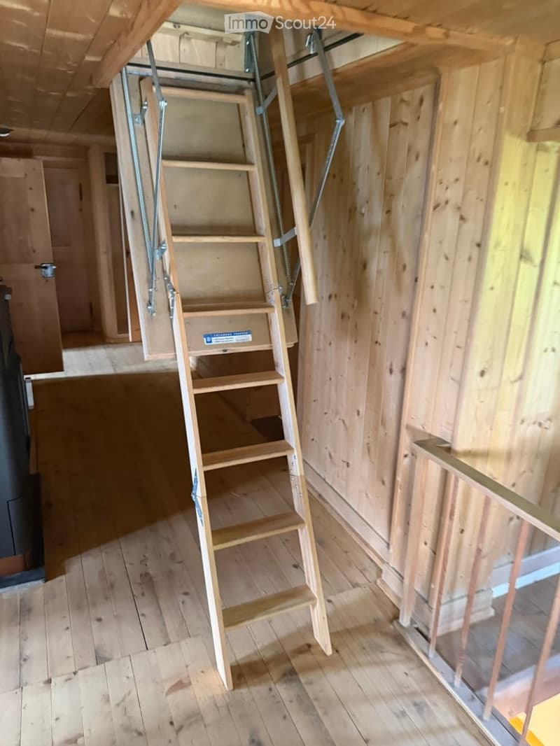 Empty room with wooden walls and floor, featuring a wooden ladder leading to an attic space.