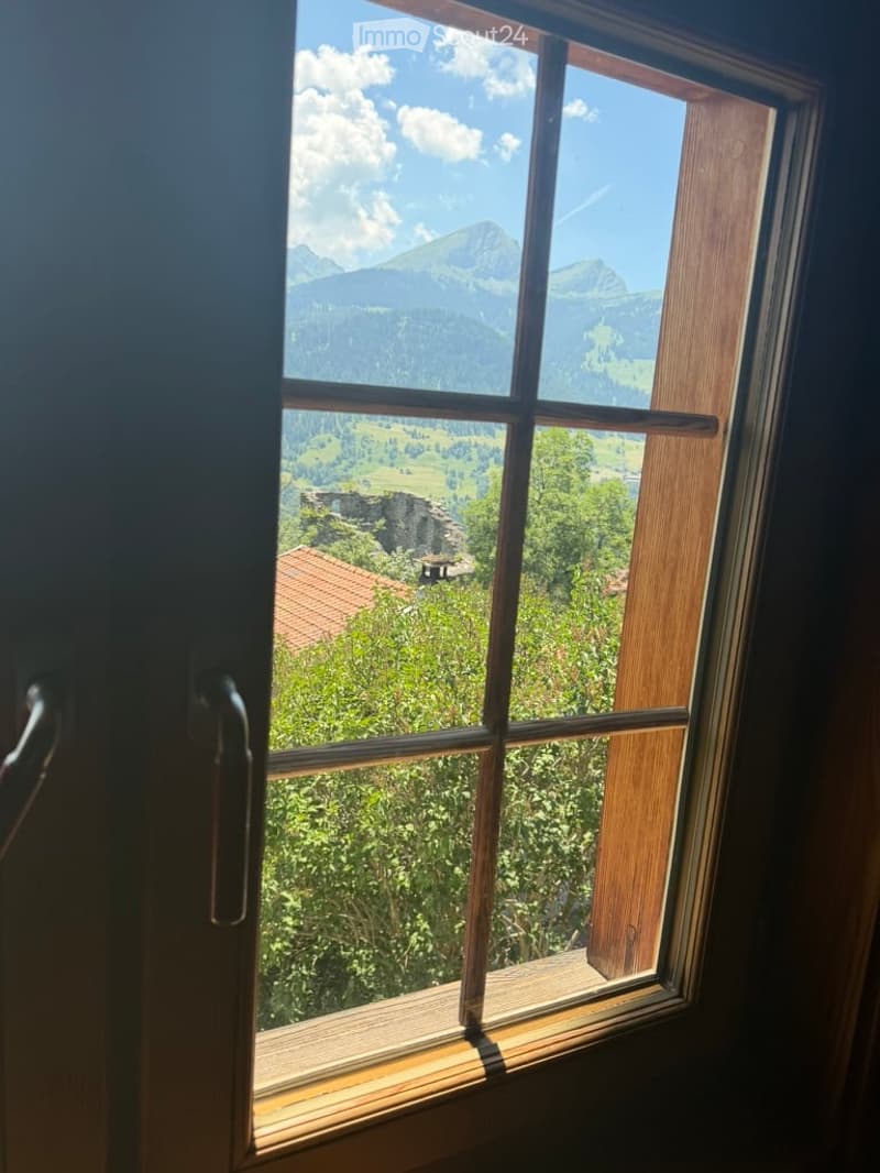 A window with wooden frame and sill, offering a scenic view of mountains, trees, and a small building in the distance.