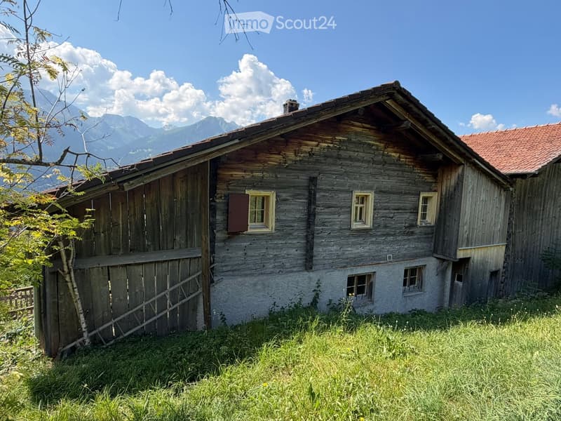 A rustic wooden house with a red-tiled roof, surrounded by grassy terrain and mountains in the background.