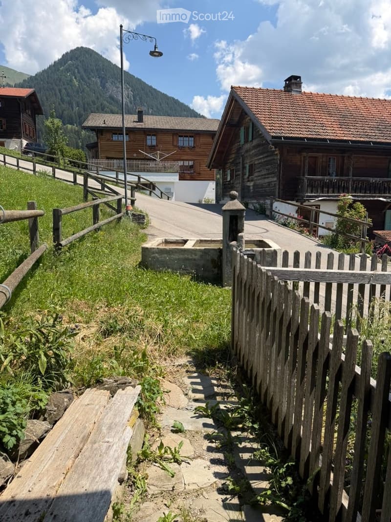 A picturesque village scene with wooden houses, a wooden fence, and a mountain backdrop under a clear blue sky.