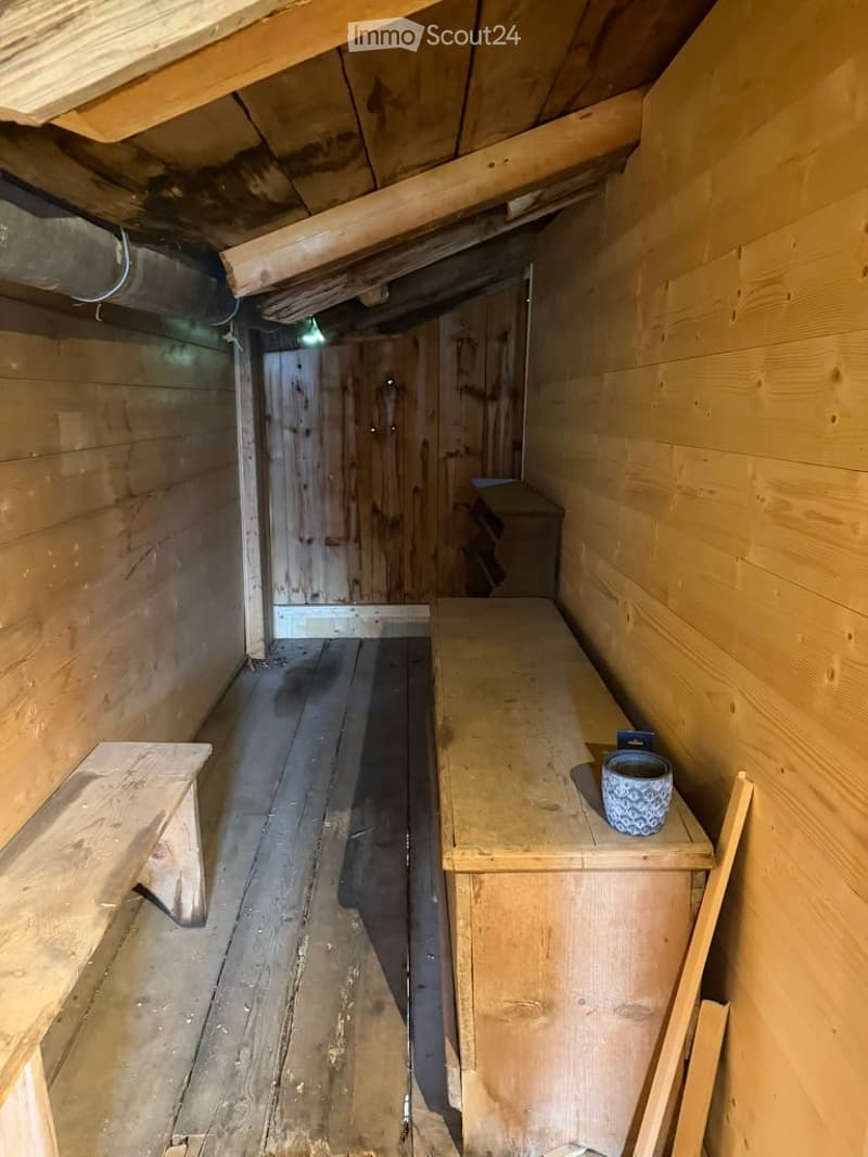 A small, rustic wooden room with exposed wooden beams and a wooden floor. The room features a wooden bench, a small wooden table, and a wooden door leading to another space.
