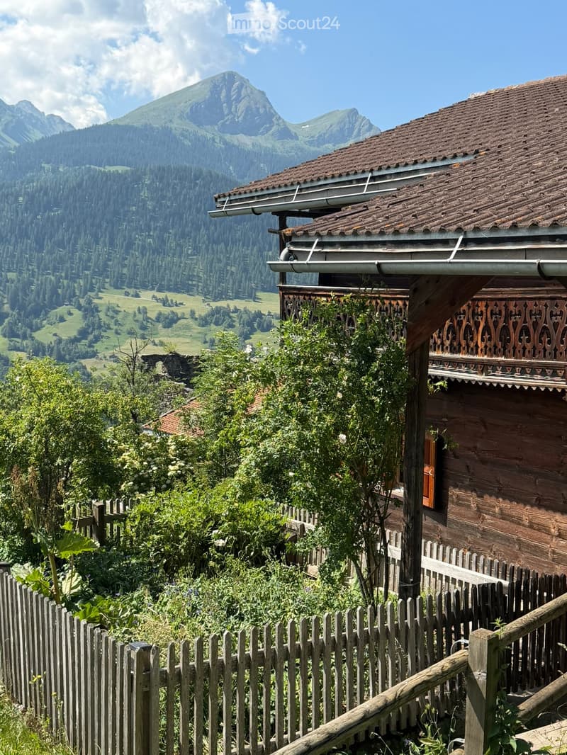 Traditional wooden house with a sloped roof and balcony, surrounded by a wooden fence and lush greenery, with a scenic mountain view in the background.