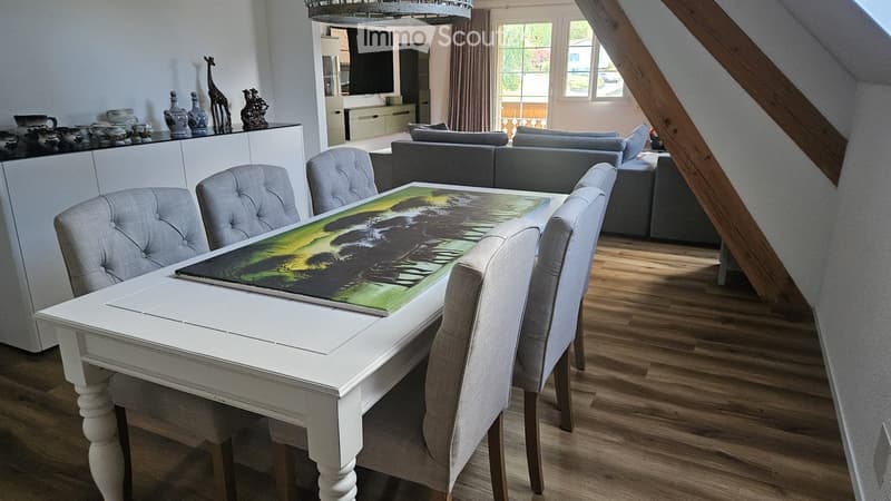 Dining area with a white wooden table, four grey upholstered chairs, and a large artwork on the table. A grey sectional sofa is visible in the background, along with a television mounted on the wall and decorative vases on a white cabinet.