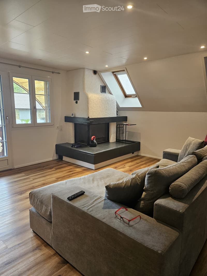 Modern living room with a grey sectional sofa, fireplace, wooden floor, and large windows. A remote control and glasses are placed on the sofa.