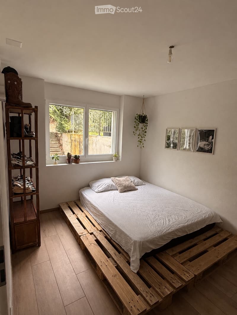 Cozy bedroom with a wooden pallet bed, white bedding, a wooden shelf with shoes and storage, and a window with potted plants on the sill.