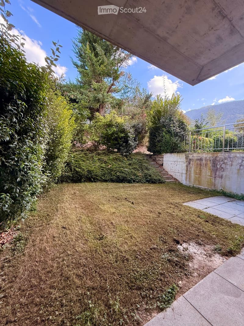 A small, slightly overgrown garden area with dry grass, concrete steps, and a metal railing leading to a higher level. The area is bordered by bushes and trees, with a clear blue sky and distant mountains visible.