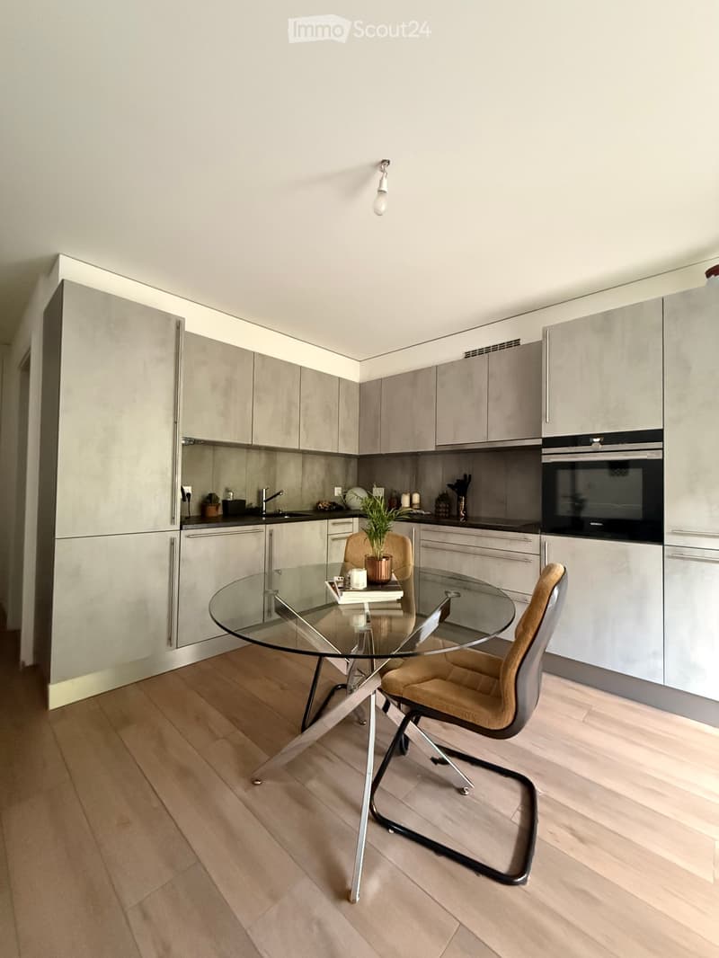 Modern kitchen with grey cabinetry, a glass dining table with chairs, and a built-in oven. The space features wooden flooring and a sleek design.