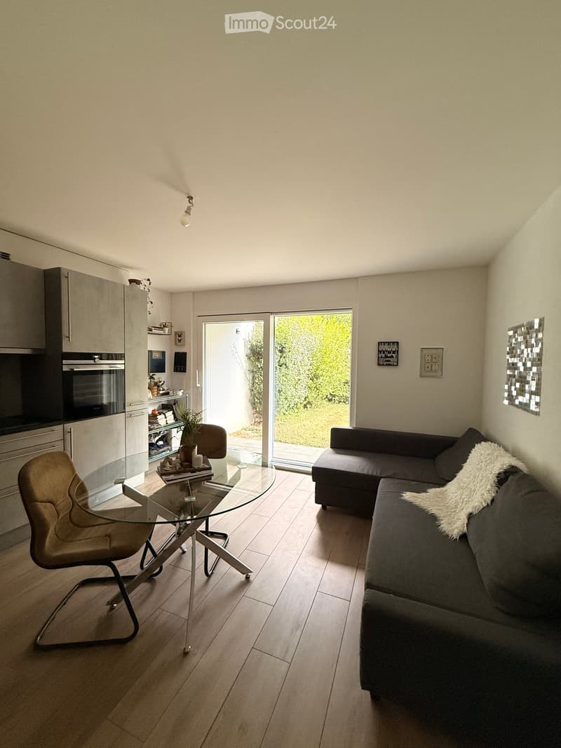 Modern living room with a grey sectional sofa, glass coffee table, armchair, and a small kitchen area with cabinets and an oven. The room features wooden flooring and large windows with a view of greenery.