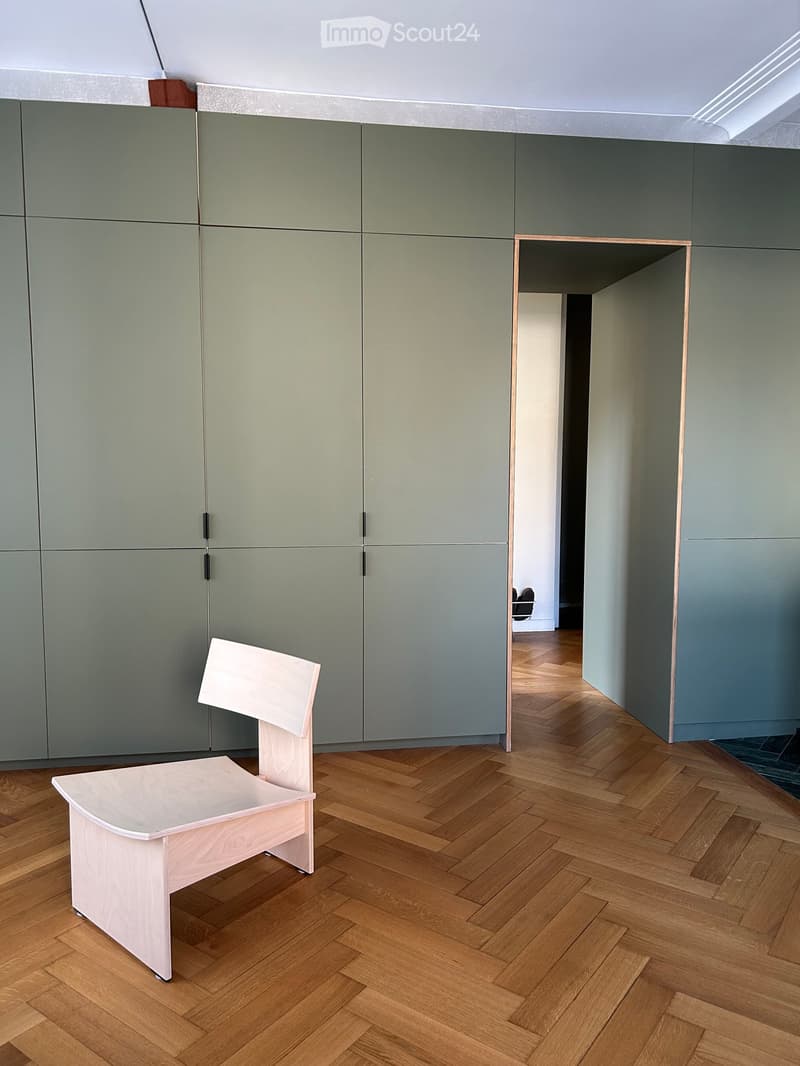A modern, minimalist room with herringbone wooden flooring, a single light-colored wooden chair, and grey built-in cabinetry.