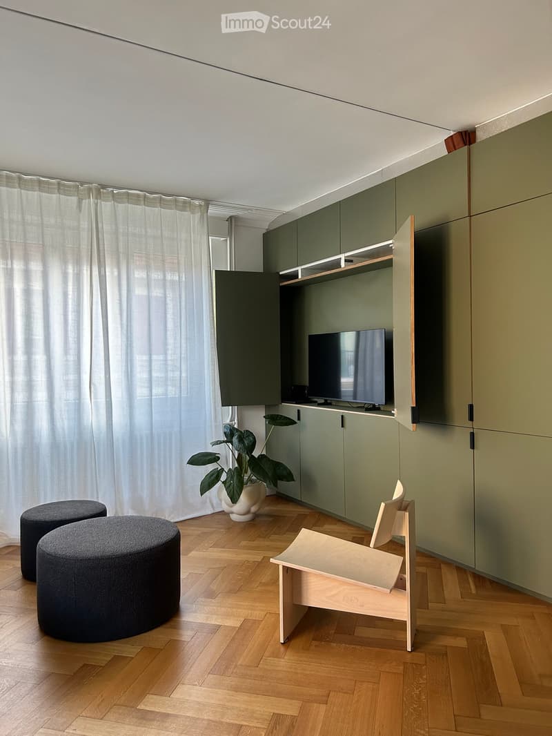 Modern living room with wooden flooring, green cabinets, a television, two round stools, a wooden chair, and a potted plant.