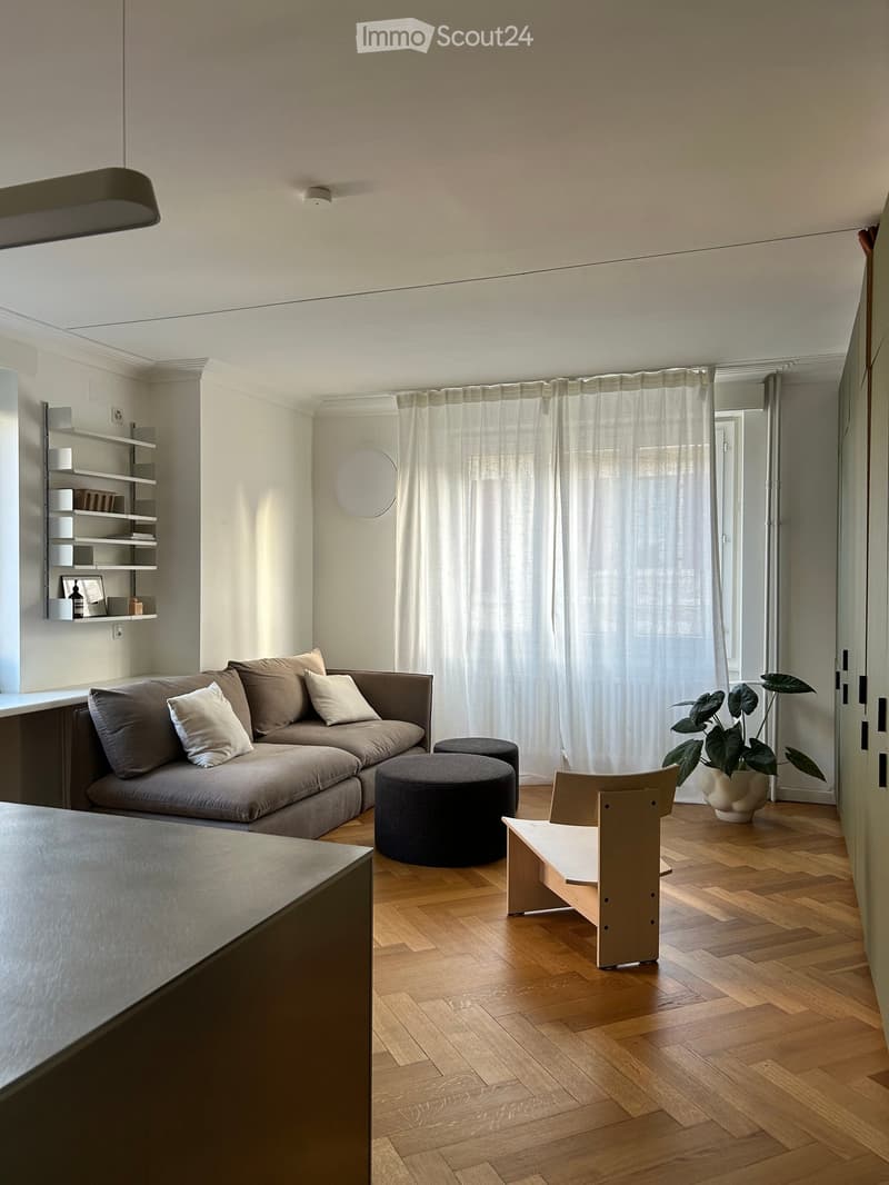 Modern living room with wooden flooring, large sectional sofa, ottoman, wooden chair, potted plant, and wall-mounted shelf.