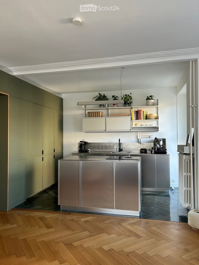 Modern kitchen with wooden herringbone flooring, stainless steel and grey cabinetry, built-in sink, coffee maker, and open shelving with plants and books.
