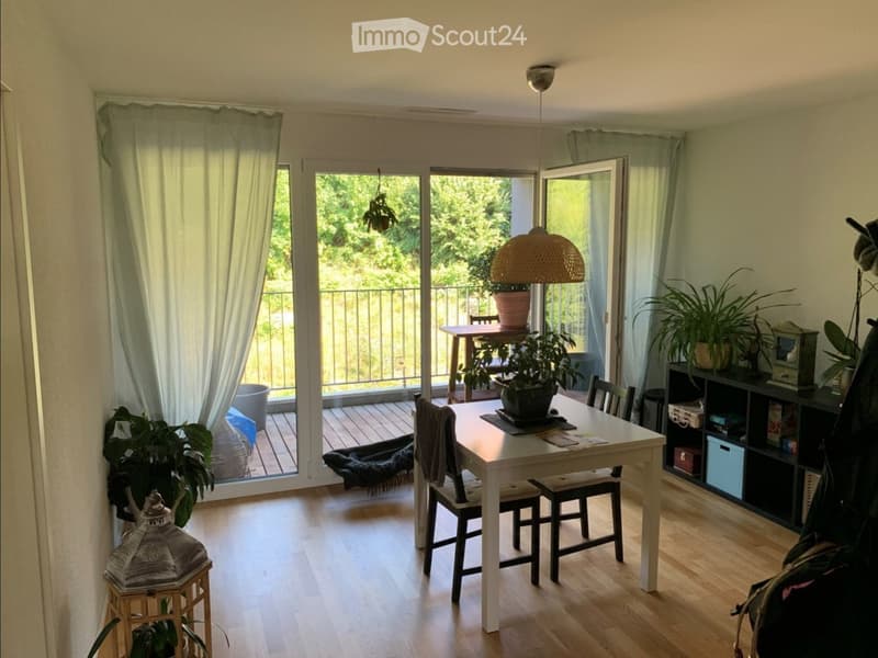 Bright dining area with a white table, four chairs, and potted plants. Large sliding glass doors open to a balcony with outdoor furniture and greenery.