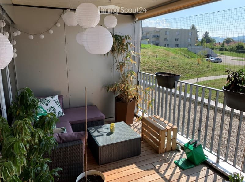 Balcony with wooden floor, sofa, small table, potted plants, and a view of a green area with buildings in the distance.