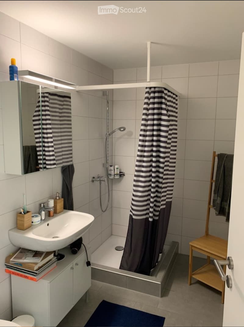 Modern bathroom with a white sink, mirror, bathtub with a black and white striped shower curtain, a small wooden stool, and a towel rack with a towel. The sink has a toothbrush holder, soap dispenser, and a small cabinet underneath holding books and other