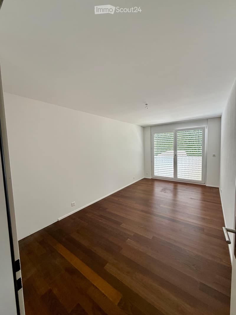 Empty room with wooden flooring and white walls, featuring a large sliding glass door with blinds leading outside.