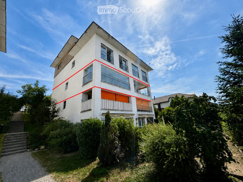 A modern, two-story house with white exterior walls, large windows, and balconies. The house is situated on a raised ground floor with a staircase leading up to it. The surrounding area includes greenery and trees.