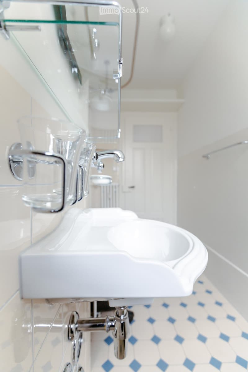 Modern bathroom with a white sink, chrome faucet, and a glass shelf above it. The floor has blue and white patterned tiles.